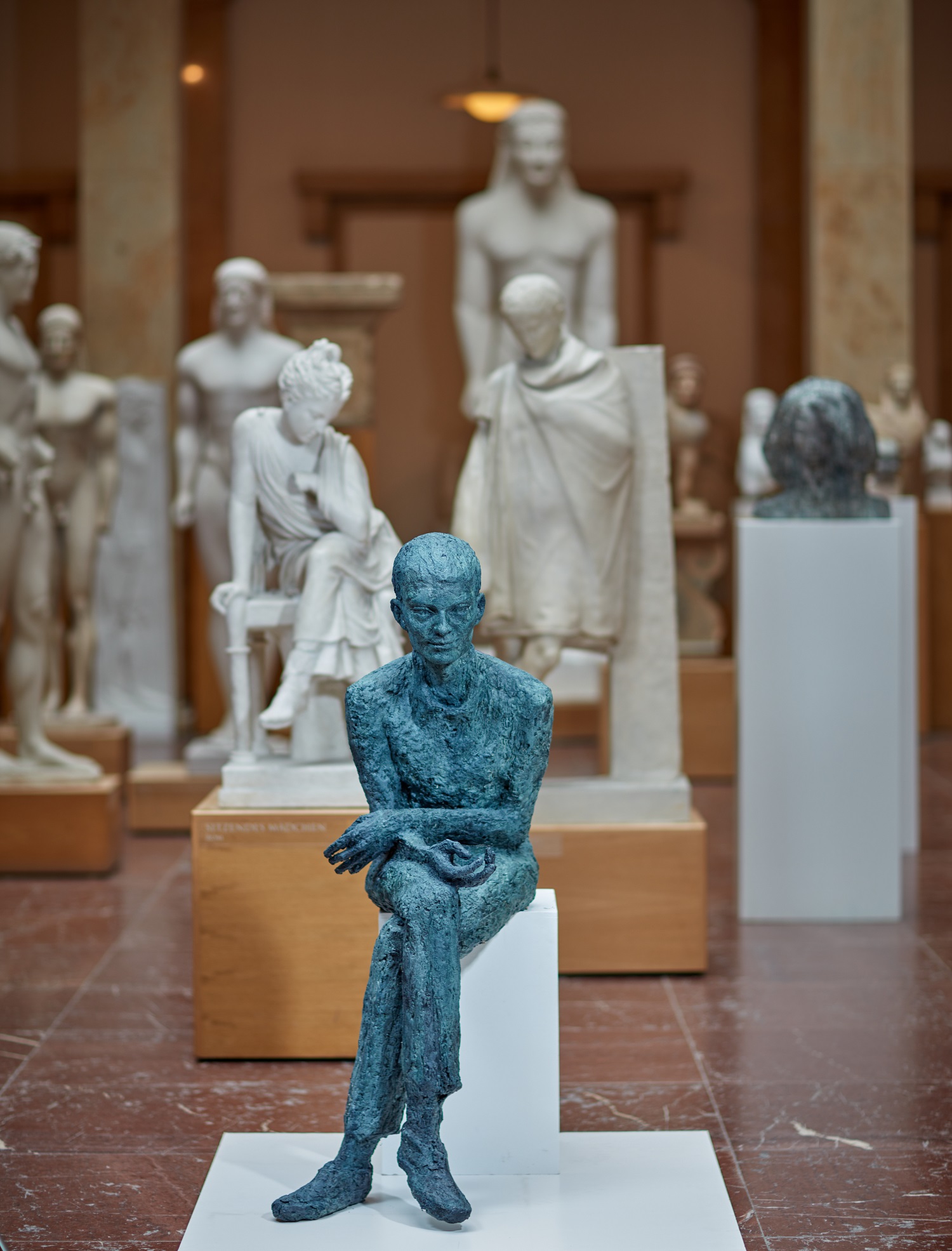 Die Figur eines jungen Mannes aus Wachs sitzt mit überschlagenen Beinen nach vorne gebeugt auf einem Sockel in den Museumsräumen.