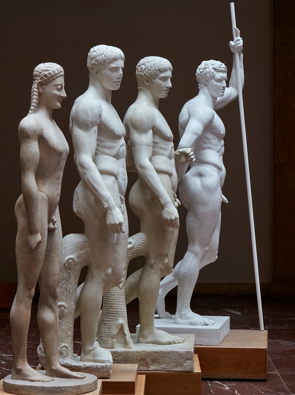 Vier nackte männliche Statuen stehen schräg nach hinten versetzt nebeneinander